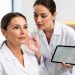 Portrait réaliste d’une femme adulte au visage naturel pendant une consultation de médecine esthétique, avec une médecin analysant ses proportions à l’aide d’une tablette dans un cabinet moderne éclairé doucement.